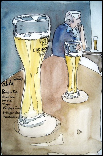 A close up of a glass of beer with a second glass on the table. In the background sits a man with his beer. 